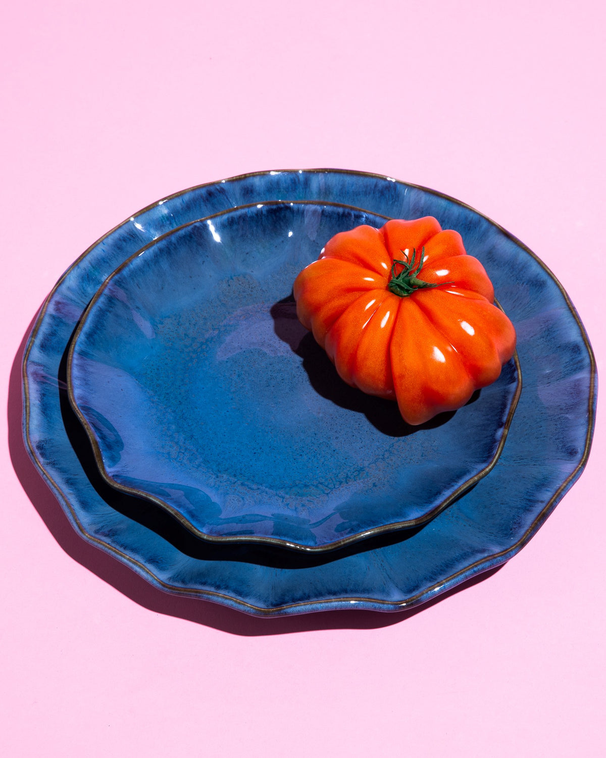 Zwei blaue Teller mit einer orangefarbenen Tomate darauf, vor einem rosa Hintergrund.