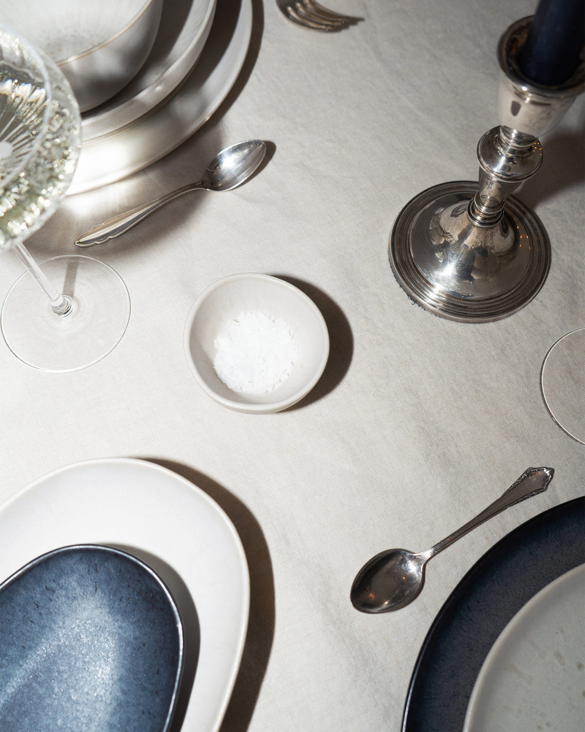 Eleganter Tisch mit Tellern, Silberbesteck, Weinglas, Kerzenhalter und einer kleinen Schale Meersalz auf weißer Tischdecke.