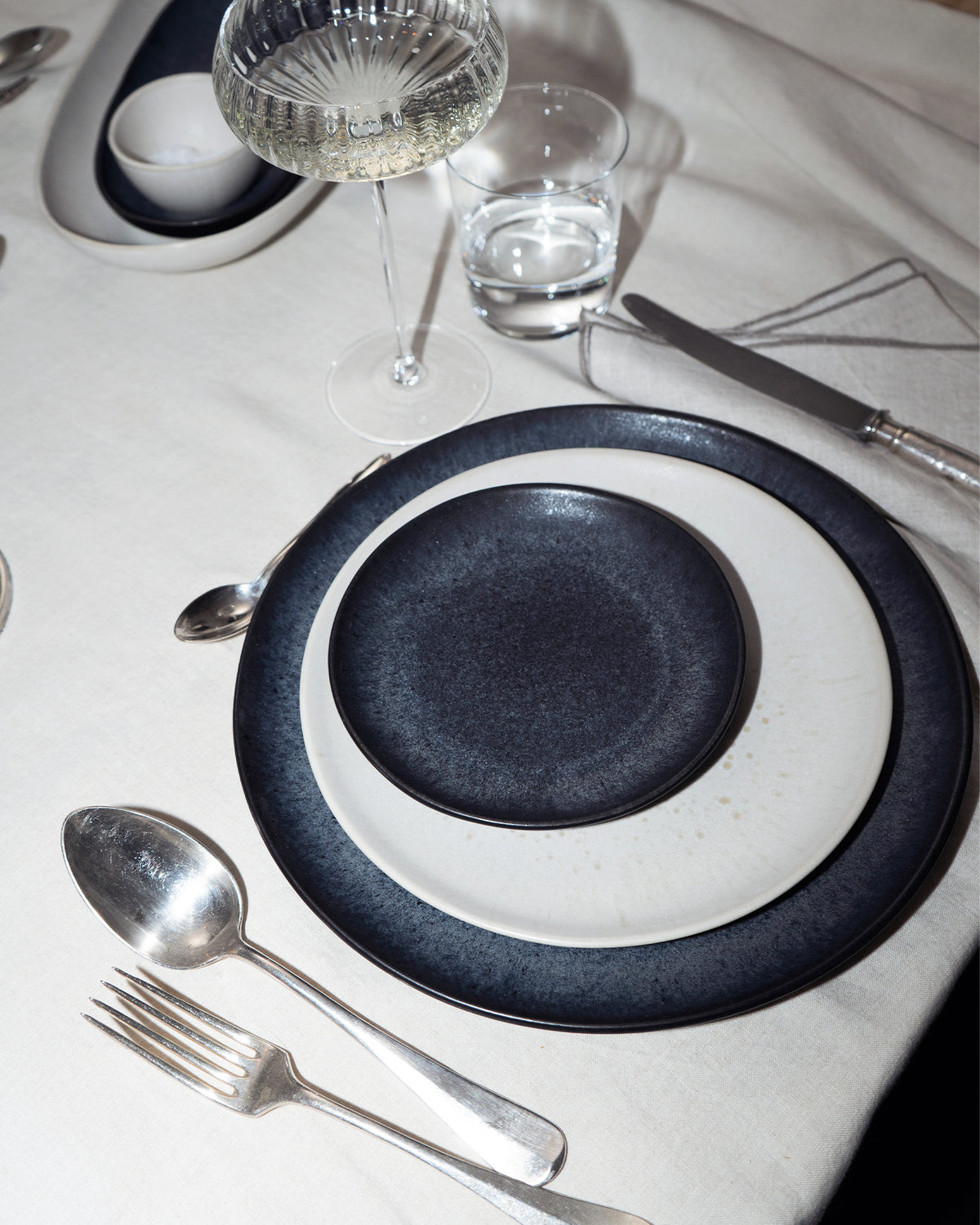 Eleganter Tisch mit gestapeltem Geschirr, Besteck, einem Weinglas und einem Wasserglas auf einer weißen Tischdecke.