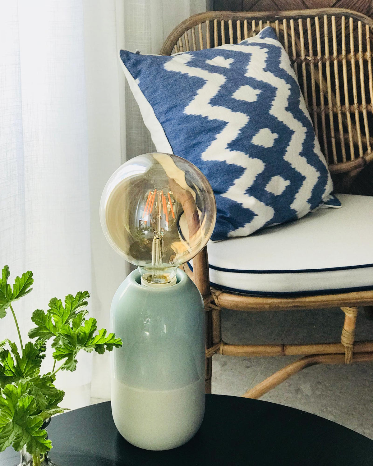 Lampe auf einem Tisch neben einem Rattanstuhl mit blau-weißem Kissen und einer Pflanze im Vordergrund.
