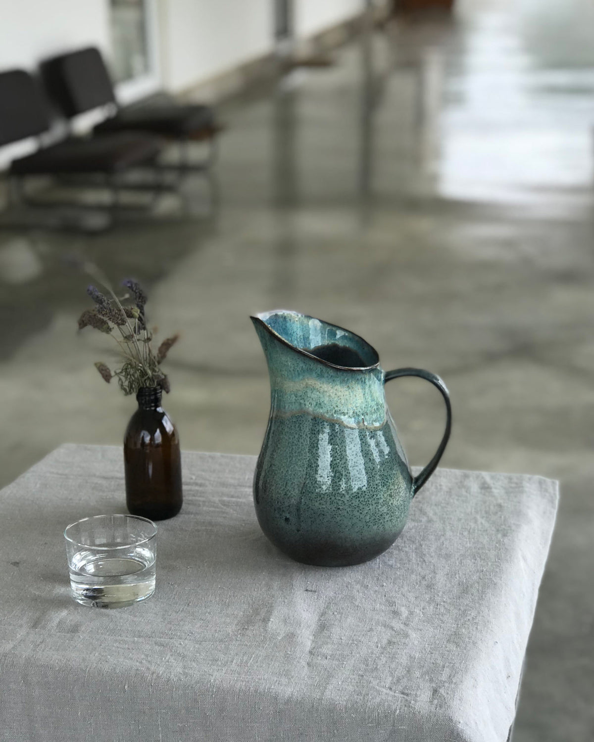 Ein grüner Krug und ein Glas Wasser stehen auf einem Tisch mit grauer Tischdecke, daneben eine Vase mit trockenen Blumen.