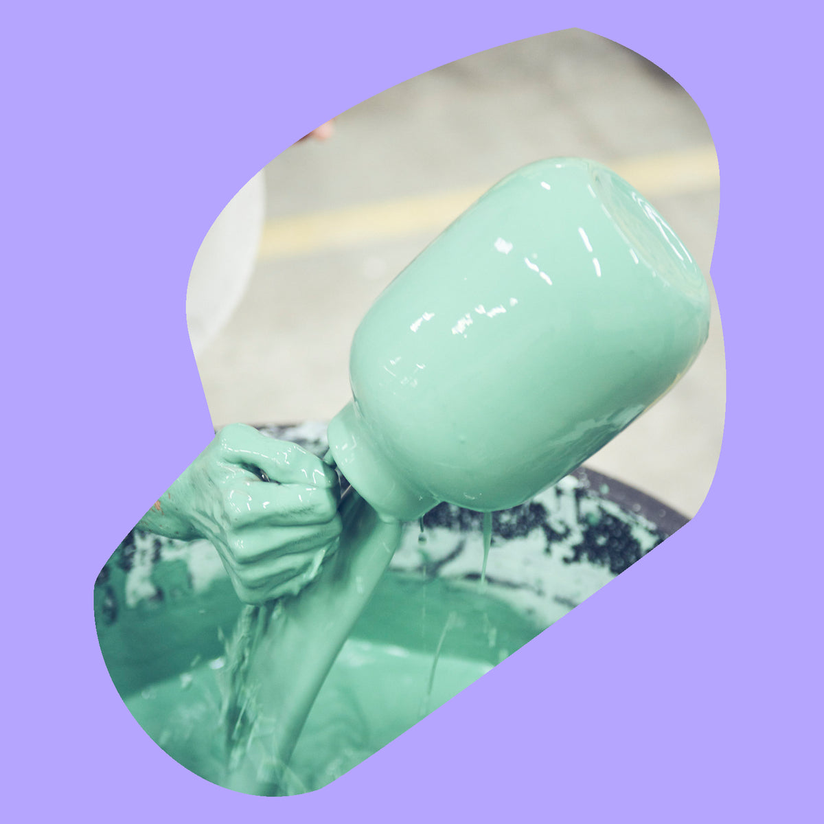Hand taucht ein blaugrünes Glasgefäß in Farbe, vor einem hellvioletten Hintergrund.
