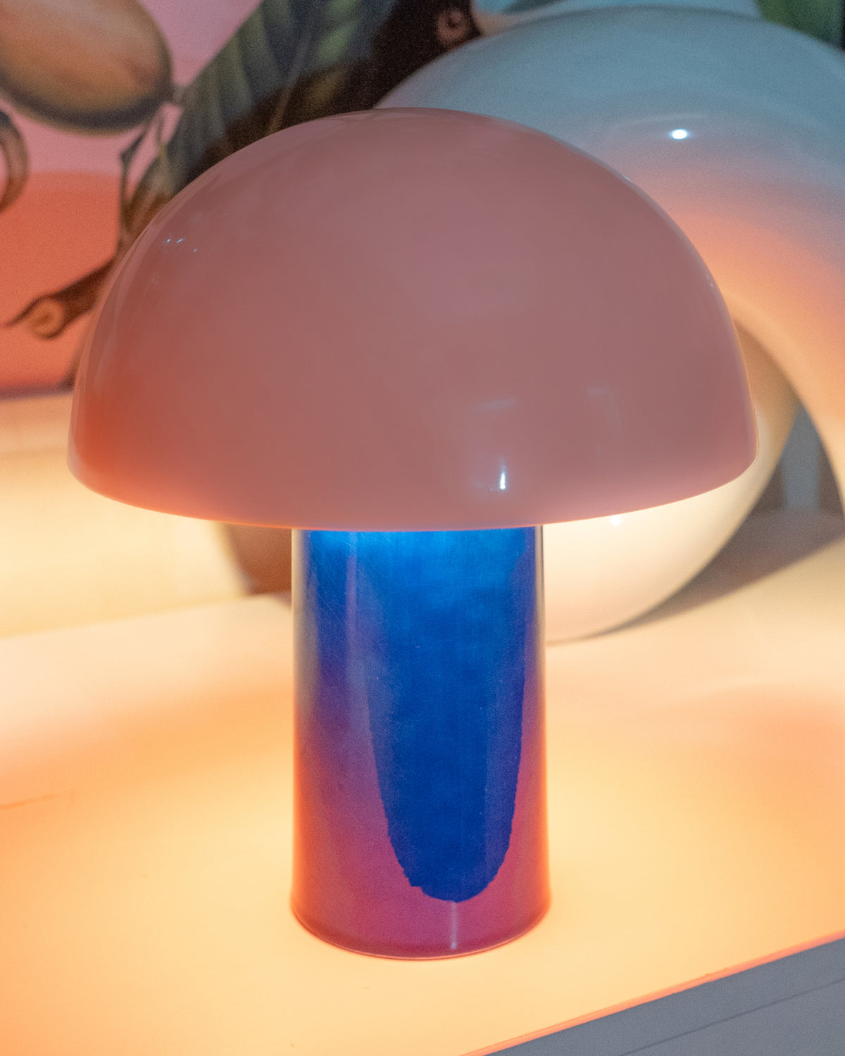 Pilzförmige Lampe mit rosafarbenem Schirm und blauem Sockel auf einem Tisch.