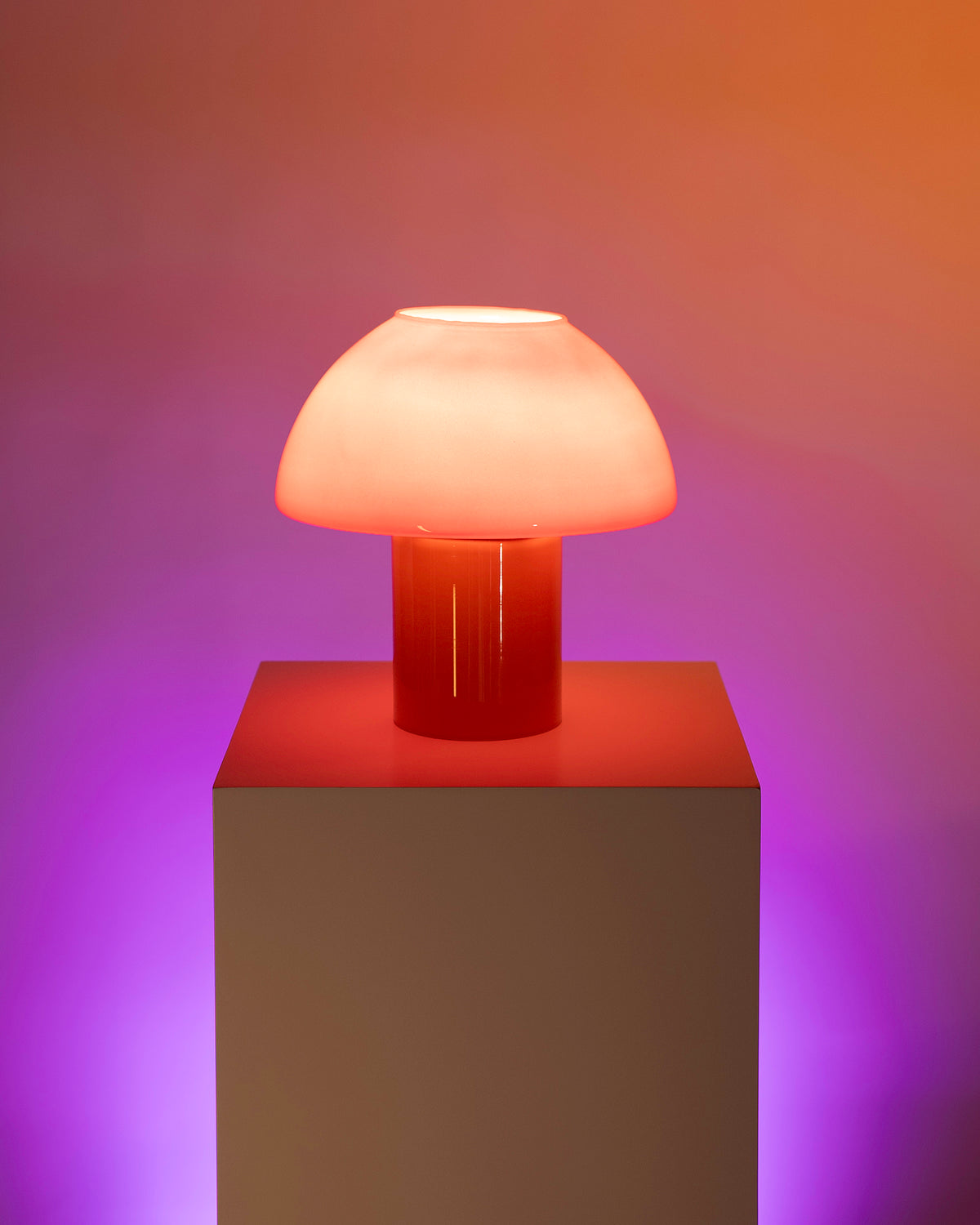 Pilzförmige, orange-rosa Lampe leuchtet auf einem Sockel vor einem farbig beleuchteten, lila-orangefarbenen Hintergrund.