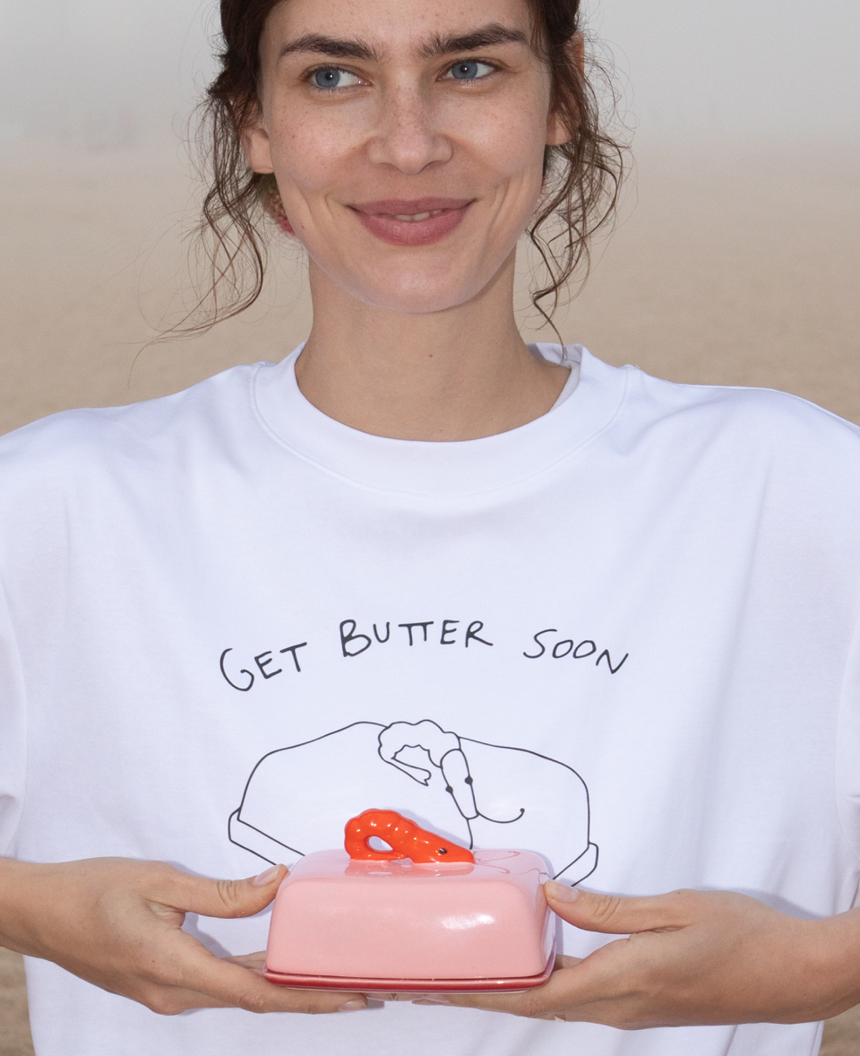 Eine Person in einem weißen T-Shirt mit der Aufschrift "Get Butter Soon" hält eine rosa Butterdose.