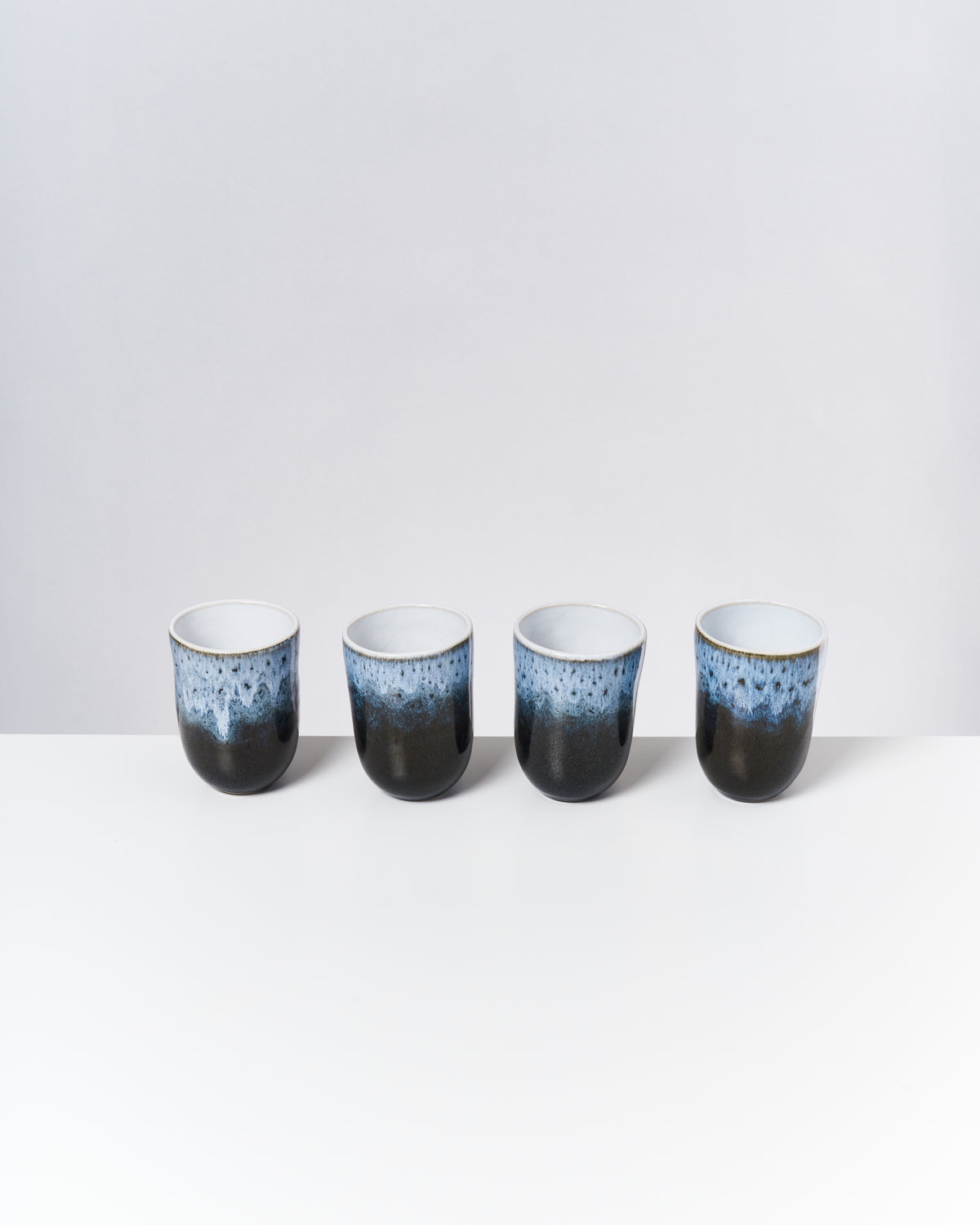 Vier schwarz-blau glasierte Keramiktassen stehen in einer Reihe auf einem weißen Tisch vor einem grauen Hintergrund.