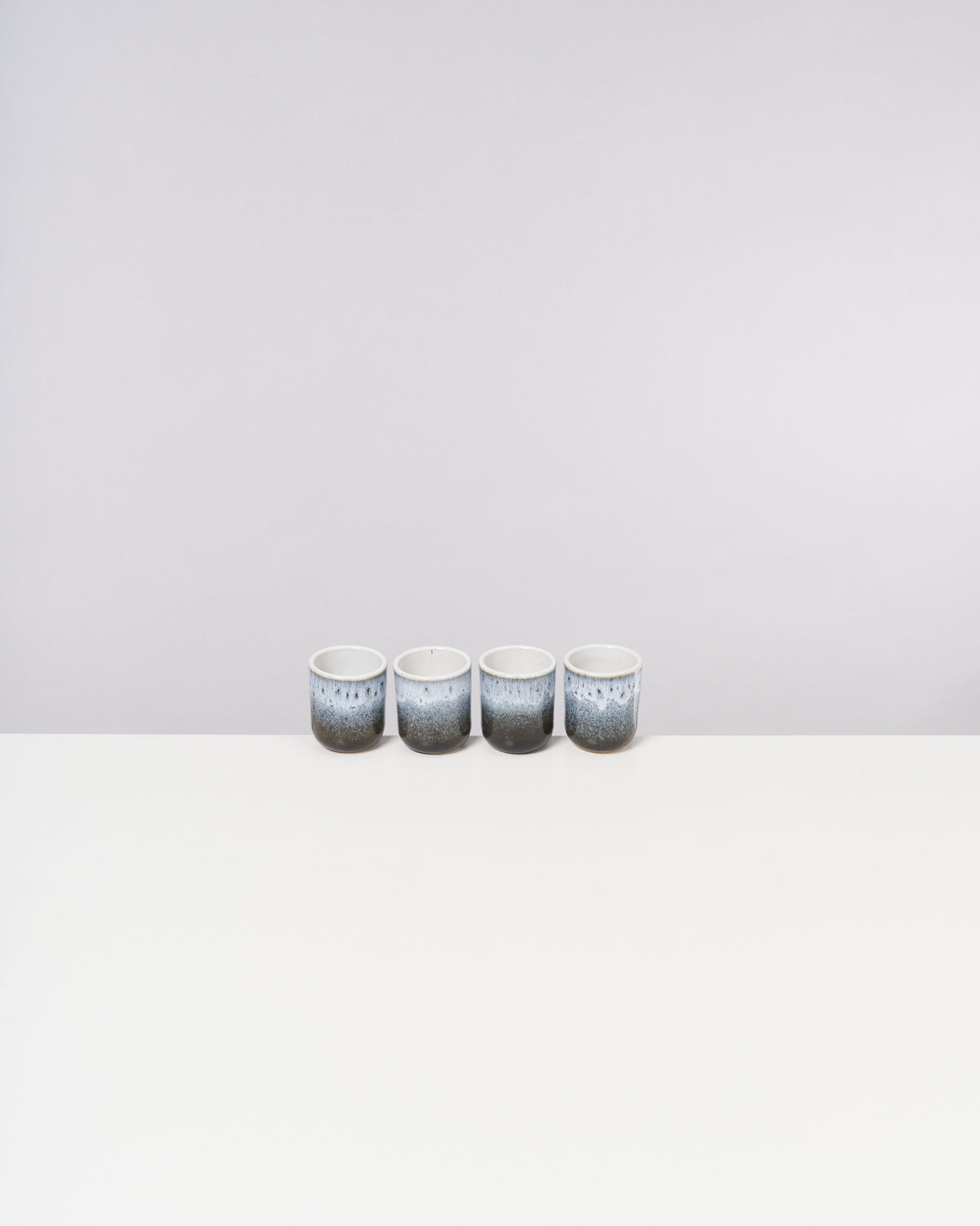 Vier Keramikbecher mit blauem Muster stehen in einer Reihe auf einem weißen Tisch vor neutralem Hintergrund.