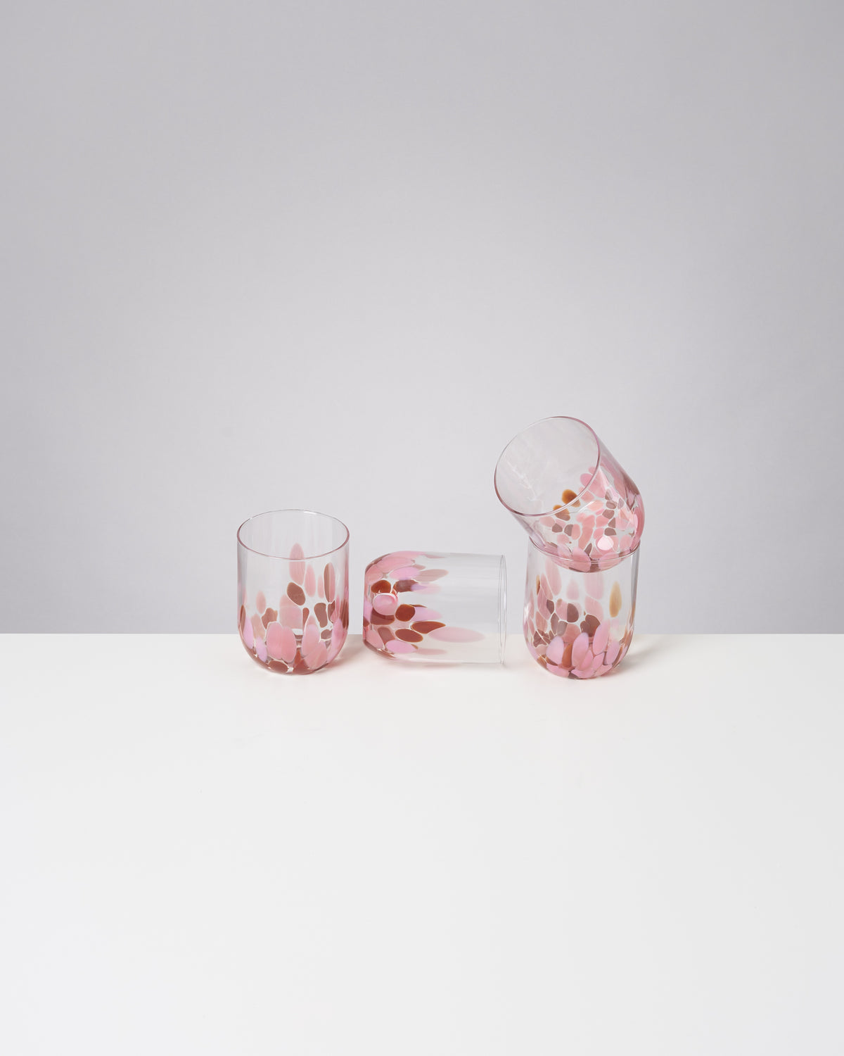 Vier gläserne Becher mit rosa und braunen Verzierungen stehen und liegen auf einem weißen Tisch vor grauem Hintergrund.