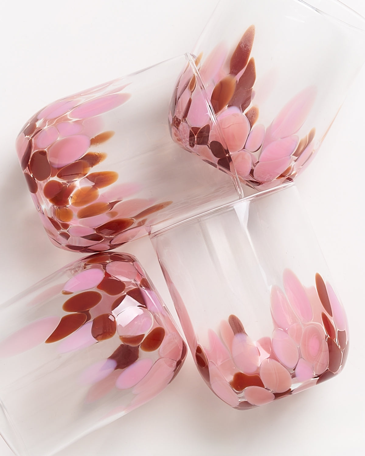 Vier Glasbecher mit rosa und braunen Mustern an den Rändern liegen durcheinander auf einer hellen Oberfläche.