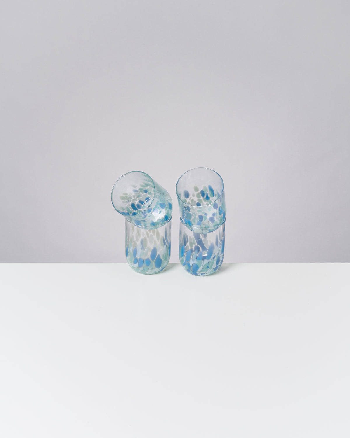 Zwei bunte Gläser mit blauen und grünen Mustern stehen auf einem weißen Tisch vor einem grauen Hintergrund.