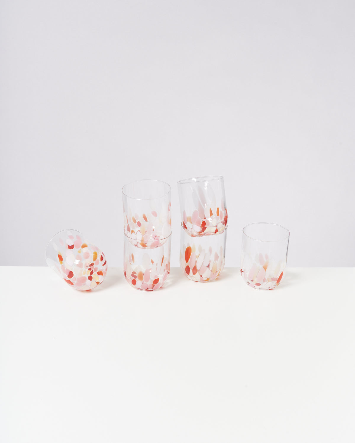 Fünf Glasbecher mit buntem Tupfenmuster stehen und liegen auf einem weißen Tisch vor hellem Hintergrund.