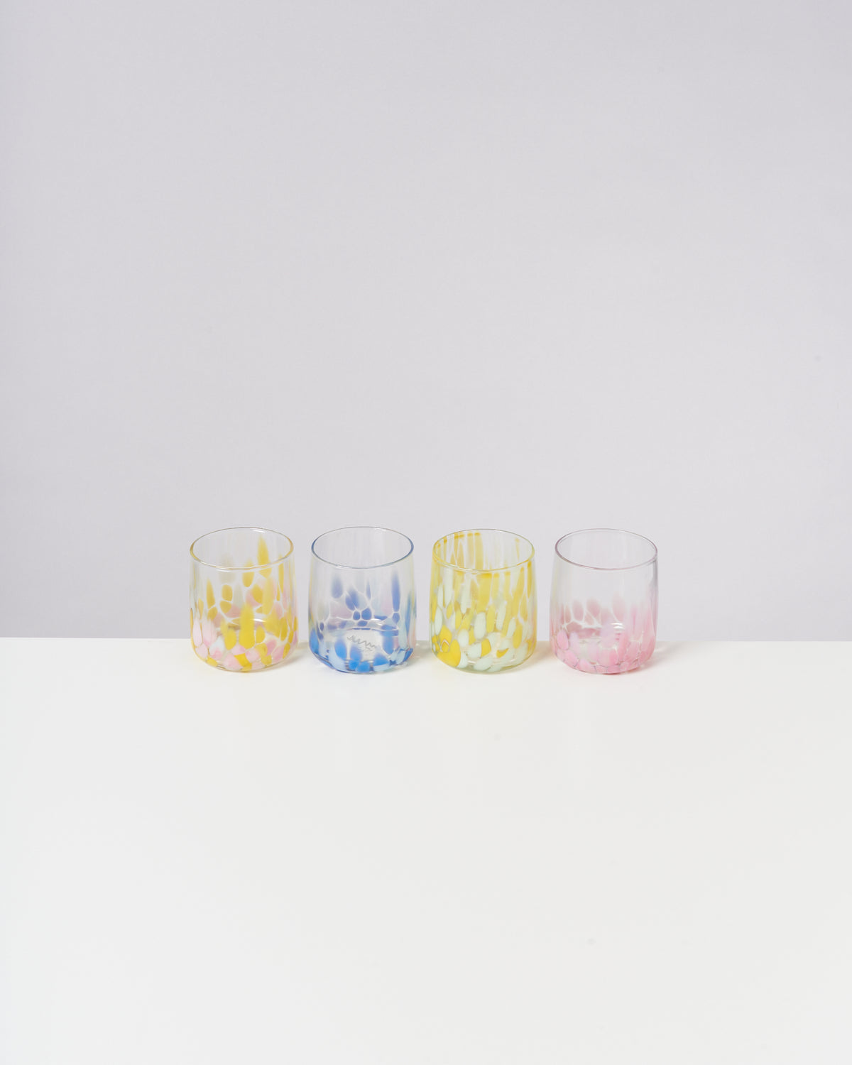 Vier bunte Gläser mit Tupfenmuster in Gelb, Blau und Rosa stehen in einer Reihe auf einem weißen Untergrund.