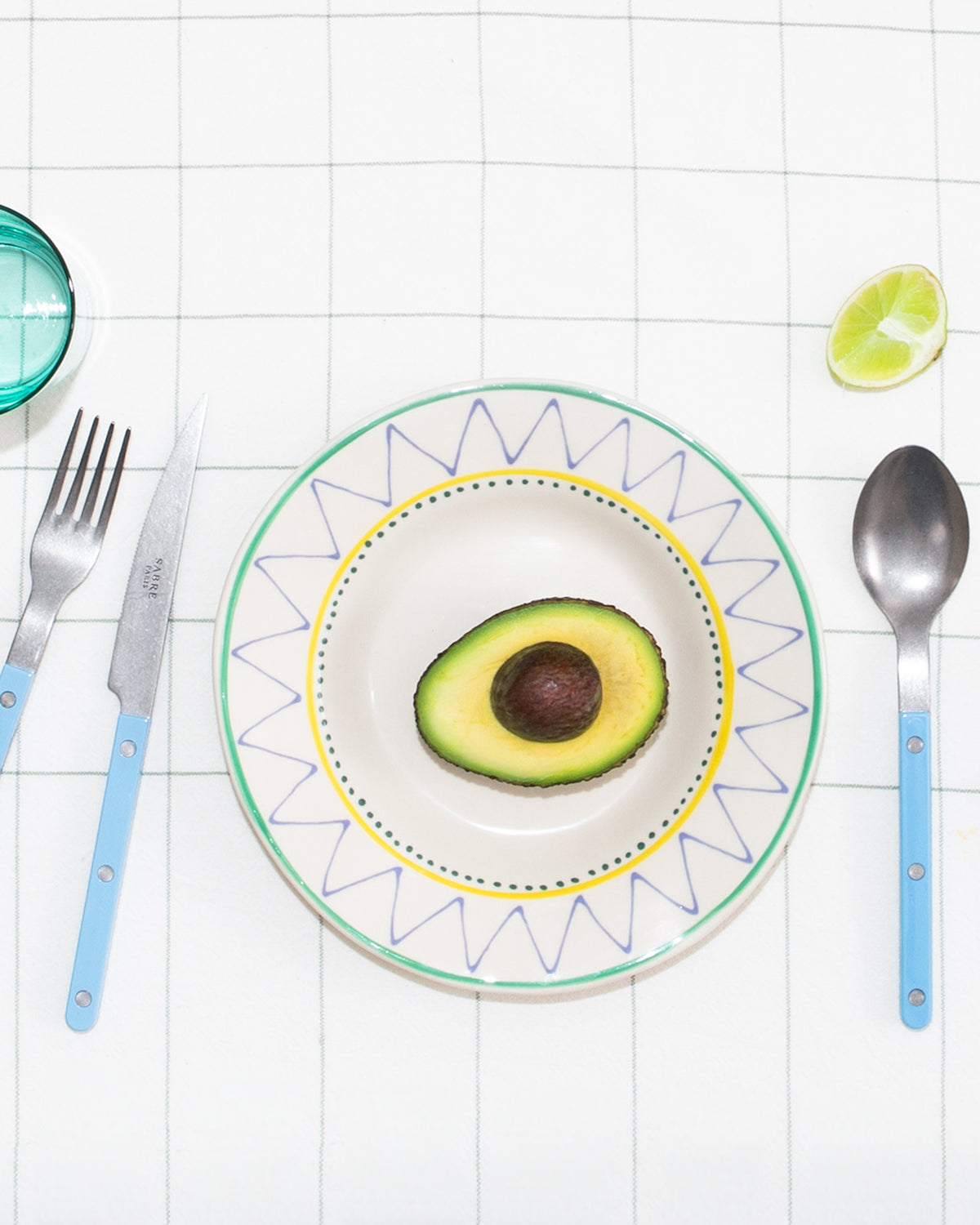 Ein Teller mit halber Avocado, Messer, Gabel und Löffel auf kariertem Tisch, daneben eine Limettenscheibe.
