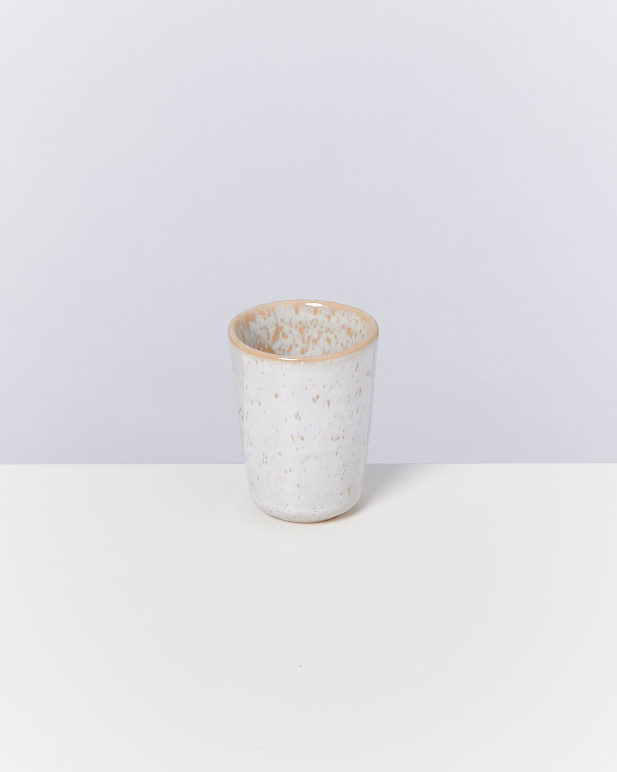 Ein weißer, gesprenkelter Keramikbecher steht auf einer weißen Oberfläche vor einem grauen Hintergrund.