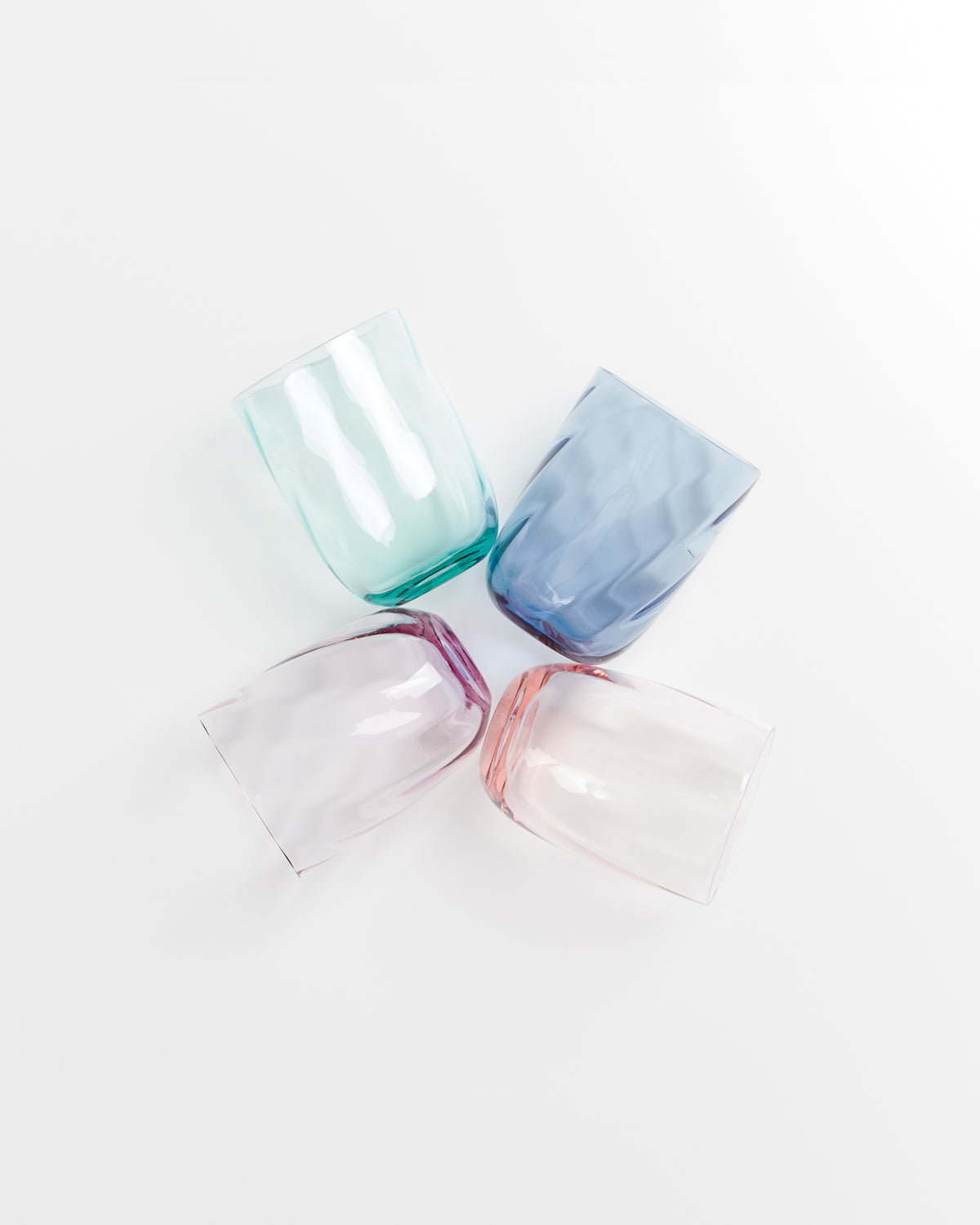 Vier bunte Gläser (grün, blau, lila, rosa) liegen auf einer weißen Oberfläche in einem Quadratmuster.