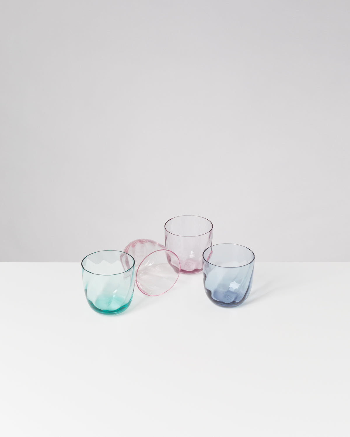 Drei bunte Trinkgläser und ein umgefallenes Glas auf weißem Hintergrund.
