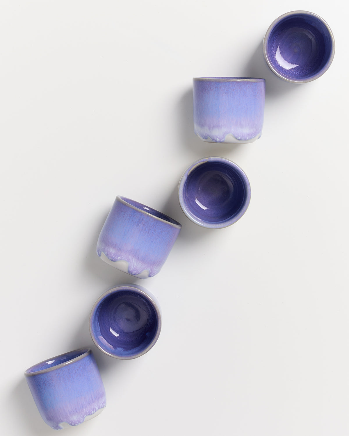 Sechs violette Tassen in einer schrägen Linie auf weißem Hintergrund angeordnet.