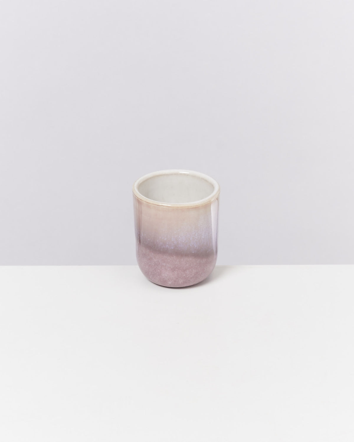 Keramiktasse mit einem Farbverlauf von Creme zu Lila, auf einer weißen Oberfläche vor grauem Hintergrund.