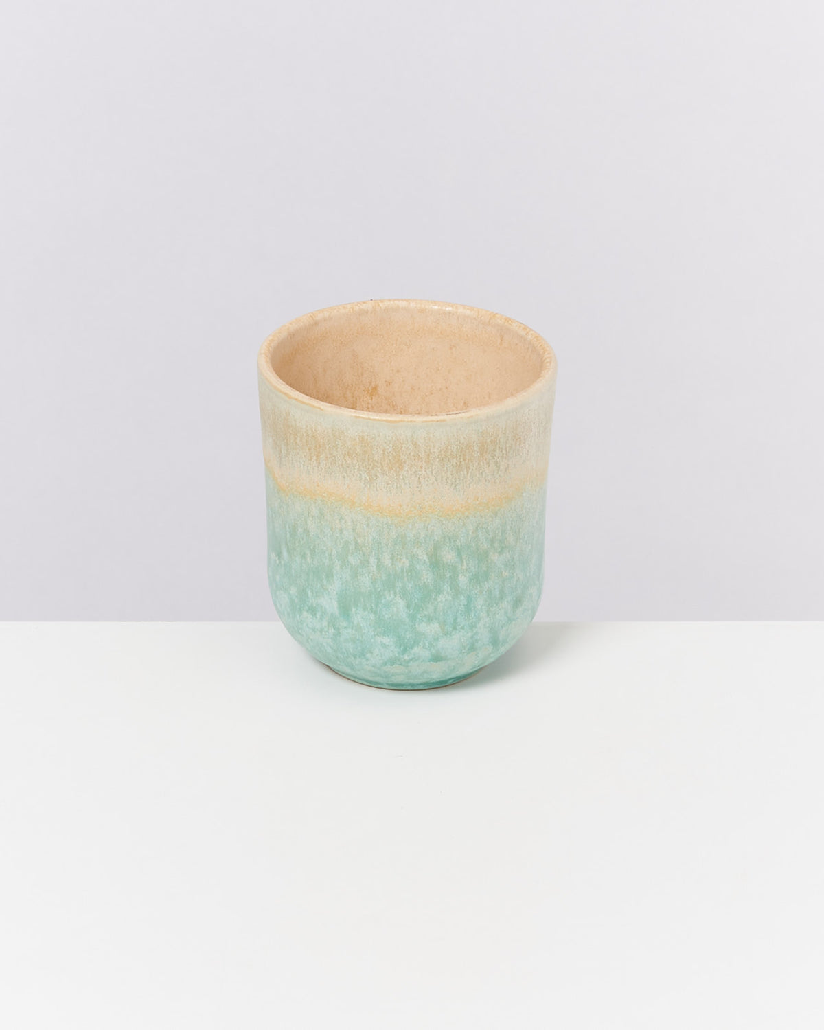 Keramiktasse mit blauer und beiger Glasur auf einem weißen Tisch vor hellem Hintergrund.