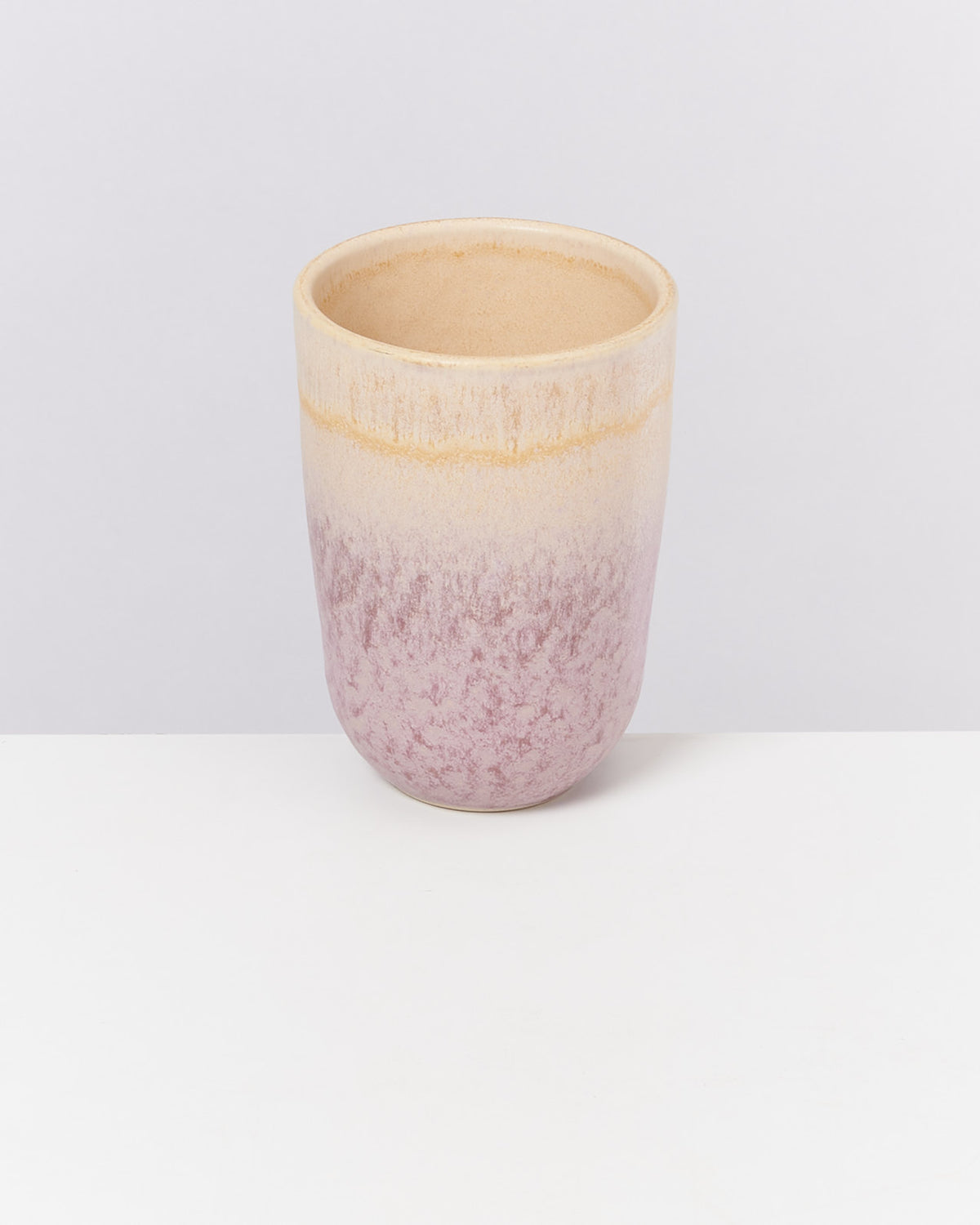 Bunt glasierter Keramikbecher mit einem Farbverlauf von oben nach unten, von cremefarben zu rosa-violett, auf weißem Tisch.