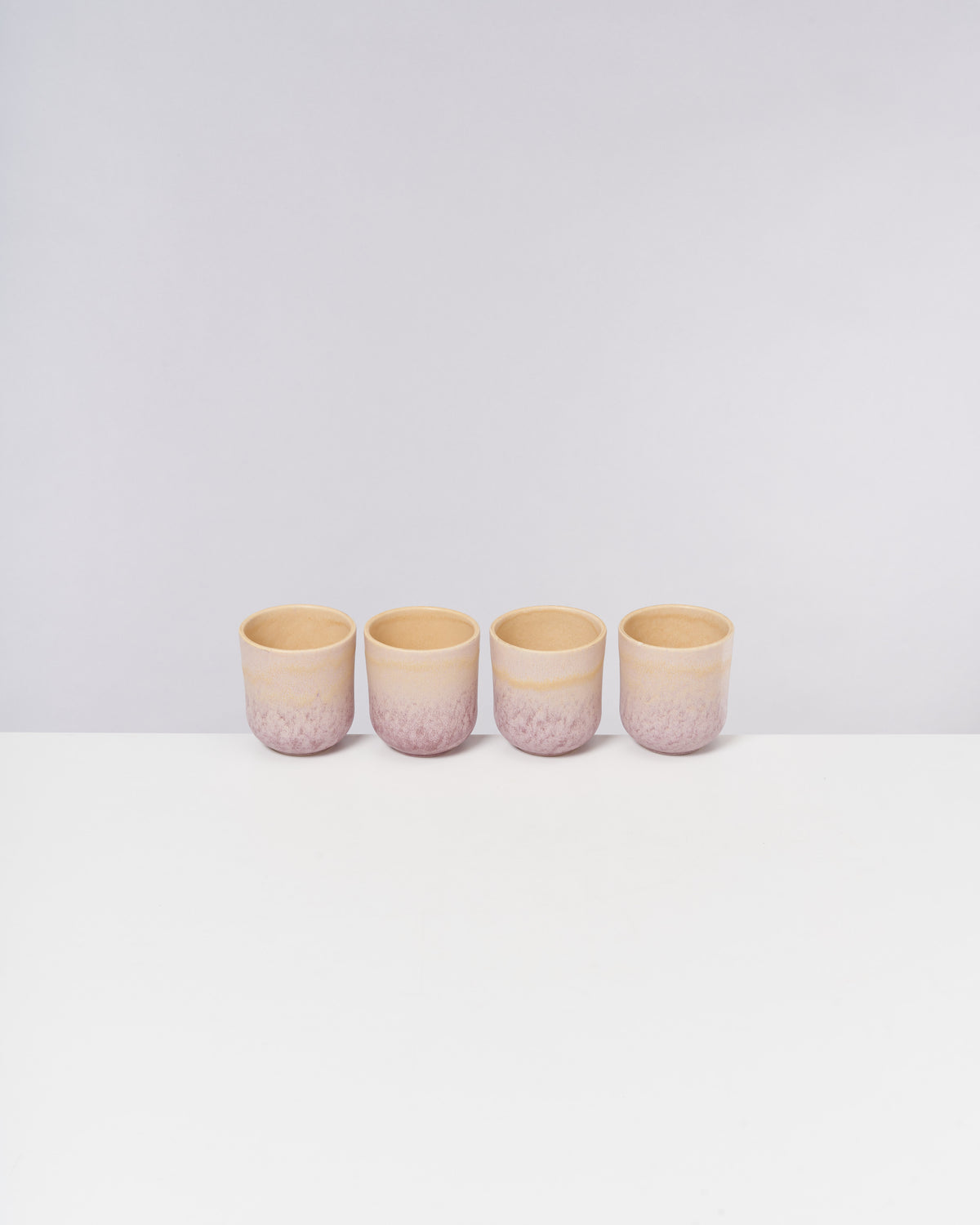 Vier beige-rosafarbene Keramikbecher stehen nebeneinander auf einer weißen Oberfläche vor grauem Hintergrund.