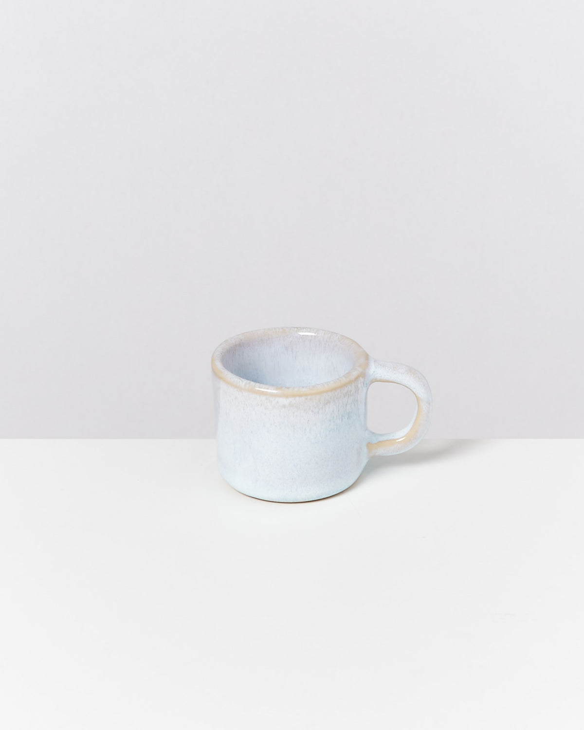 Eine kleine, matte, weiße Tasse mit einem Henkel steht auf einer weißen Oberfläche.