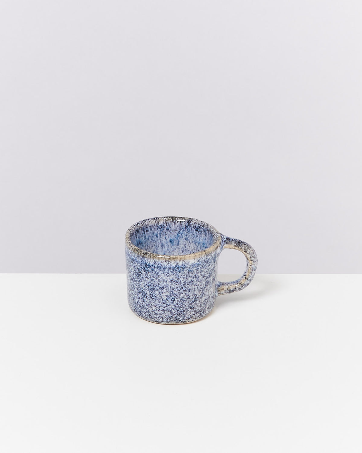 Blaue, gesprenkelte Keramiktasse mit Henkel steht auf einem weißen Tisch vor einem hellgrauen Hintergrund.