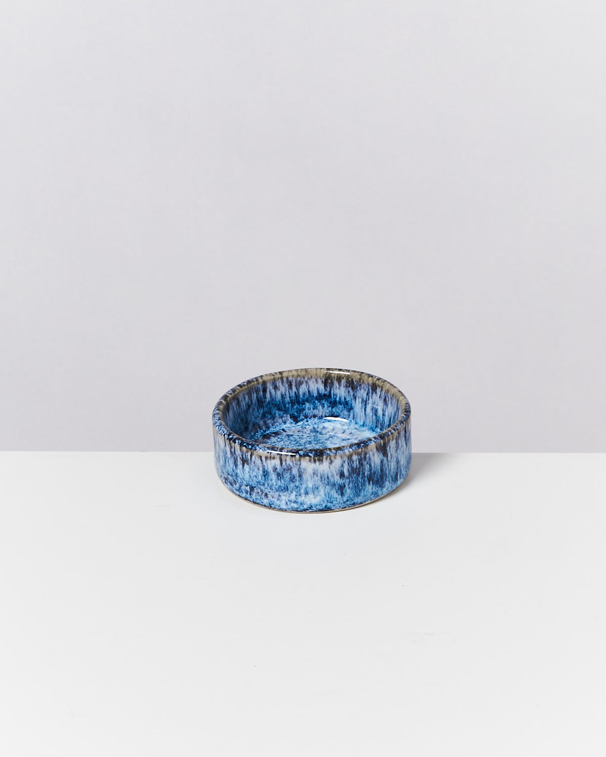 Runde, blau-graue Keramikschale mit unregelmäßigem Muster auf einer weißen Oberfläche vor grauem Hintergrund.