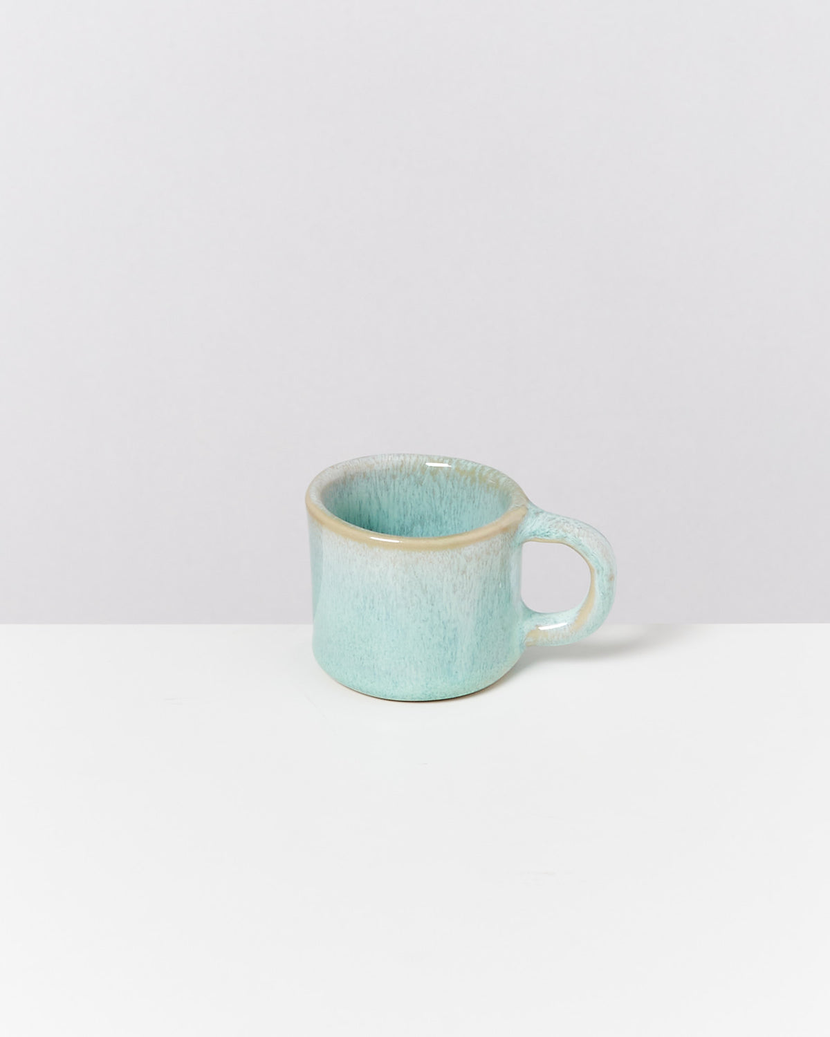 Eine hellblaue Keramiktasse mit kurzem Henkel steht auf einem weißen Tisch vor einem grauen Hintergrund.
