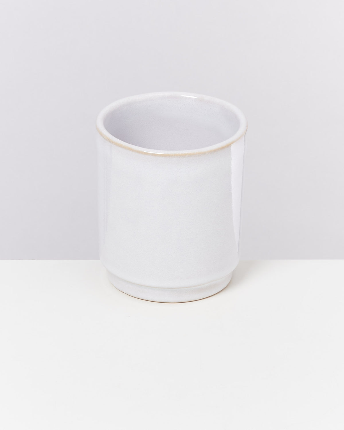 Ein weißer Keramikbecher steht auf einer weißen Oberfläche vor einem hellgrauen Hintergrund.