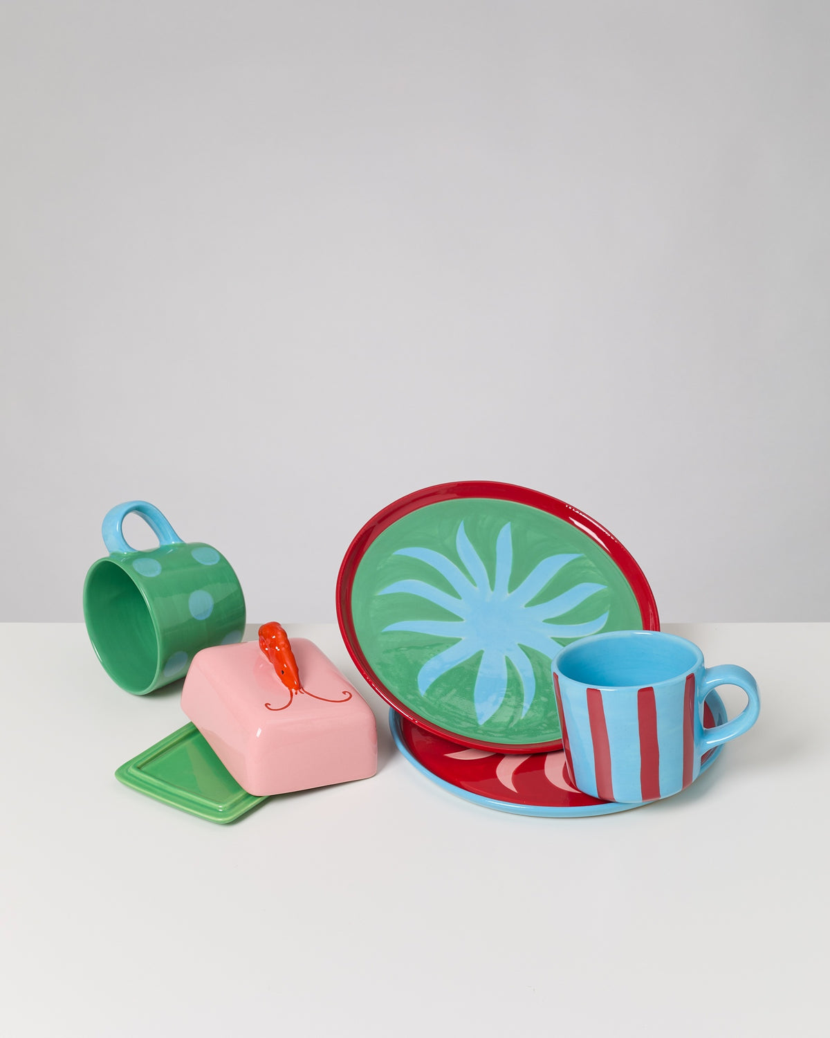 Buntes Geschirr-Set mit zwei Tassen, zwei Tellern und einer rosa Butterdose mit einem orangefarbenen Griff auf einem Tisch.