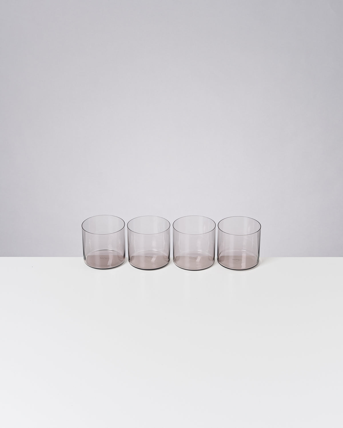 Vier leere, transparente Gläser stehen in einer Reihe auf einem weißen Tisch vor einem grauen Hintergrund.