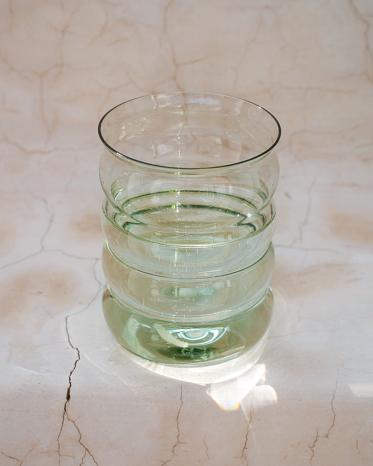 Glas mit gewellter Form steht auf einem marmorierten Untergrund und reflektiert Licht.
