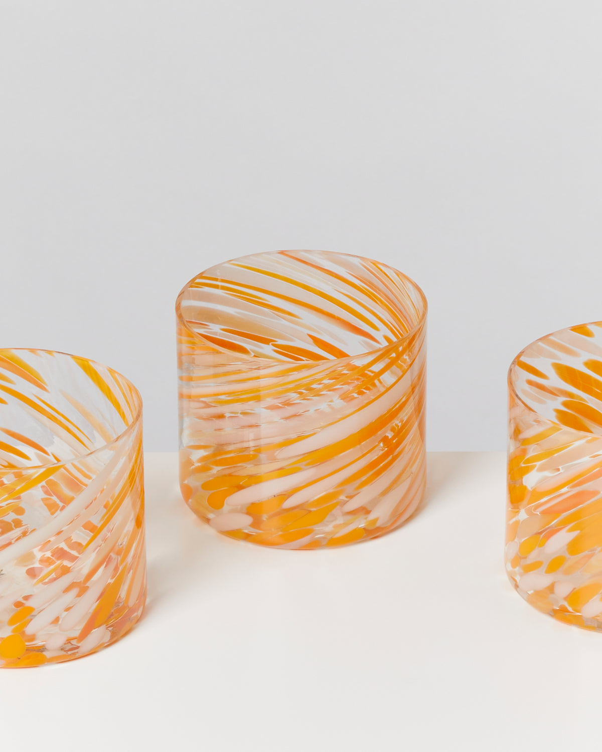 Drei gläserne Zylinder mit orange-weißen Wirbelmustern auf einer weißen Oberfläche.