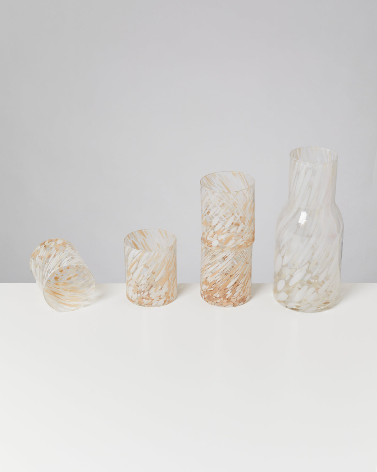 Vier Glasbecher und eine Karaffe mit weiß-goldenem Muster stehen auf einem weißen Tisch vor einem grauen Hintergrund.