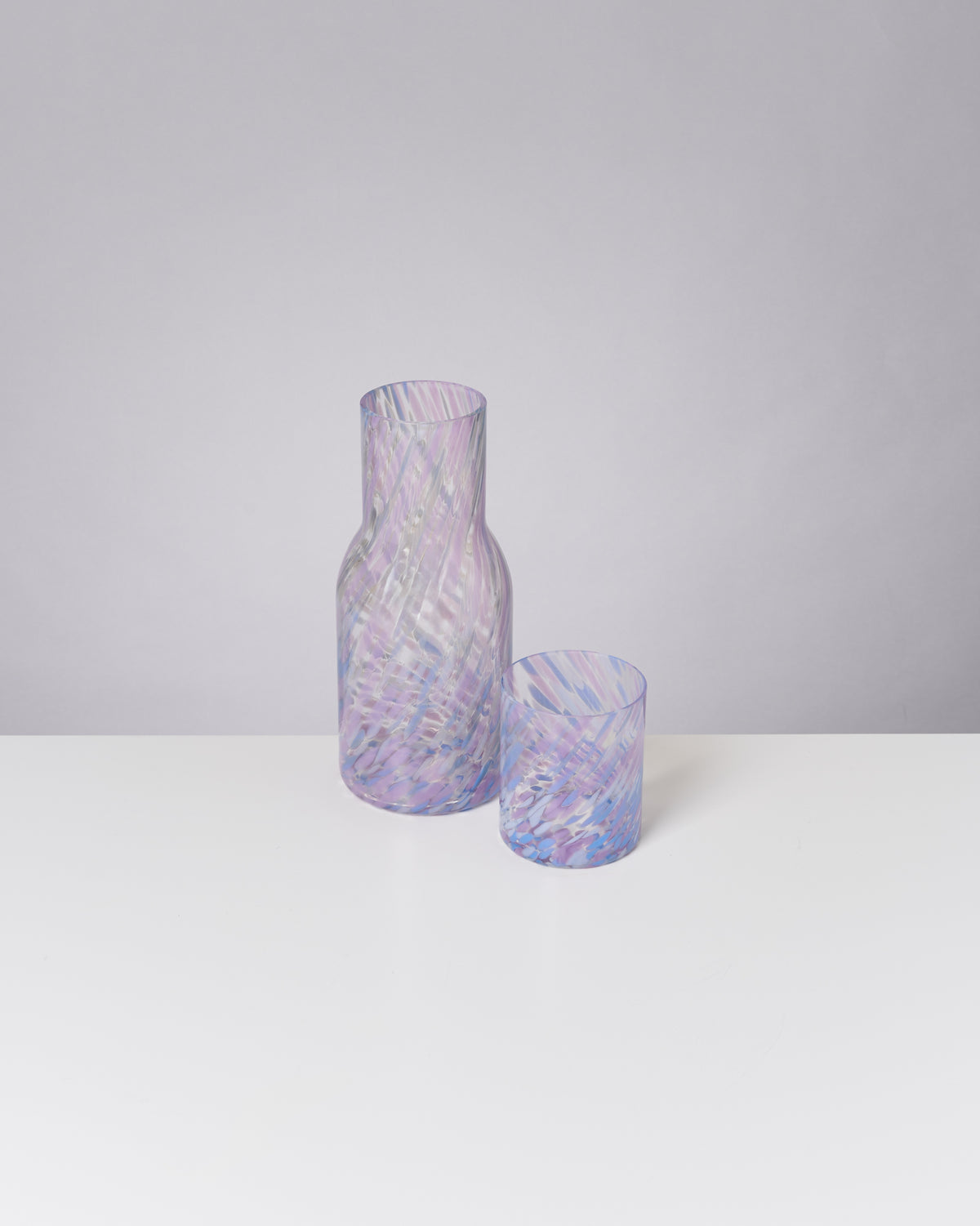 Lila und weiß gemusterte Glasflasche und -becher stehen auf einem weißen Tisch vor einem grauen Hintergrund.