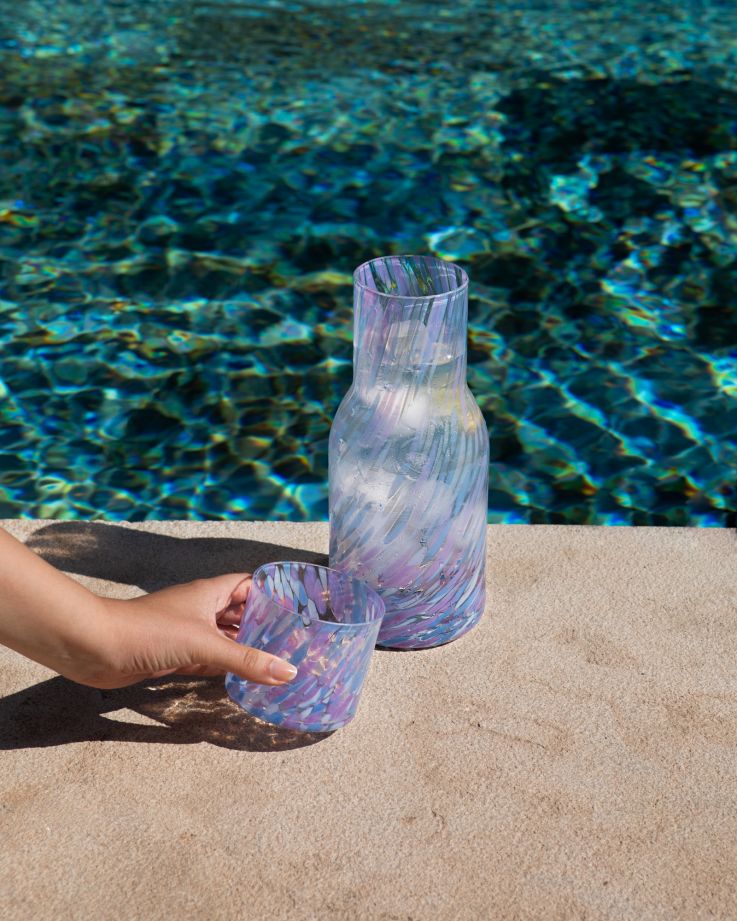 Hand greift nach einem bunten Glas neben einer passenden Karaffe am Poolrand.