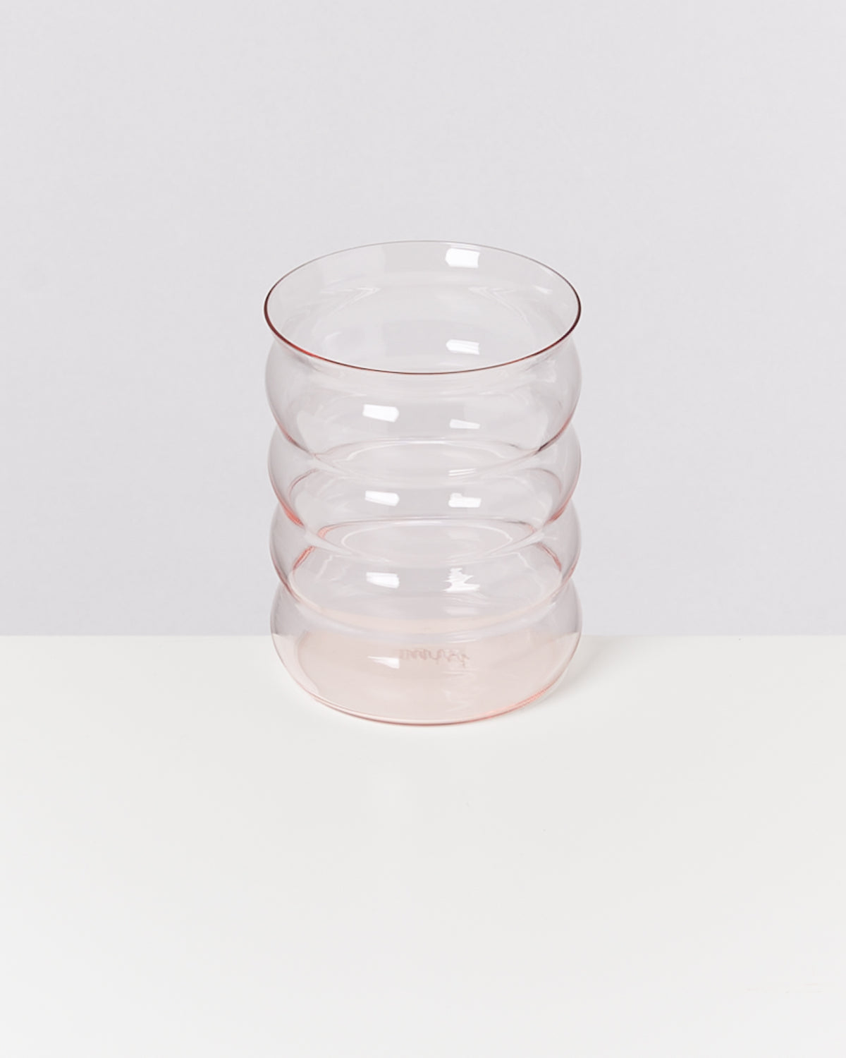 Ein transparentes Glas mit wellenförmigem Design steht auf einer weißen Oberfläche vor einem grauen Hintergrund.