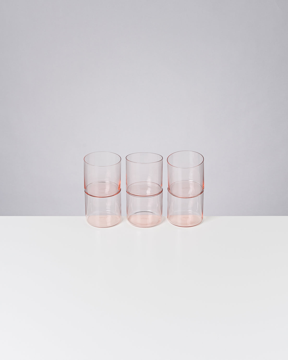 Drei transparente rosa Gläser auf einem weißen Tisch vor grauem Hintergrund.