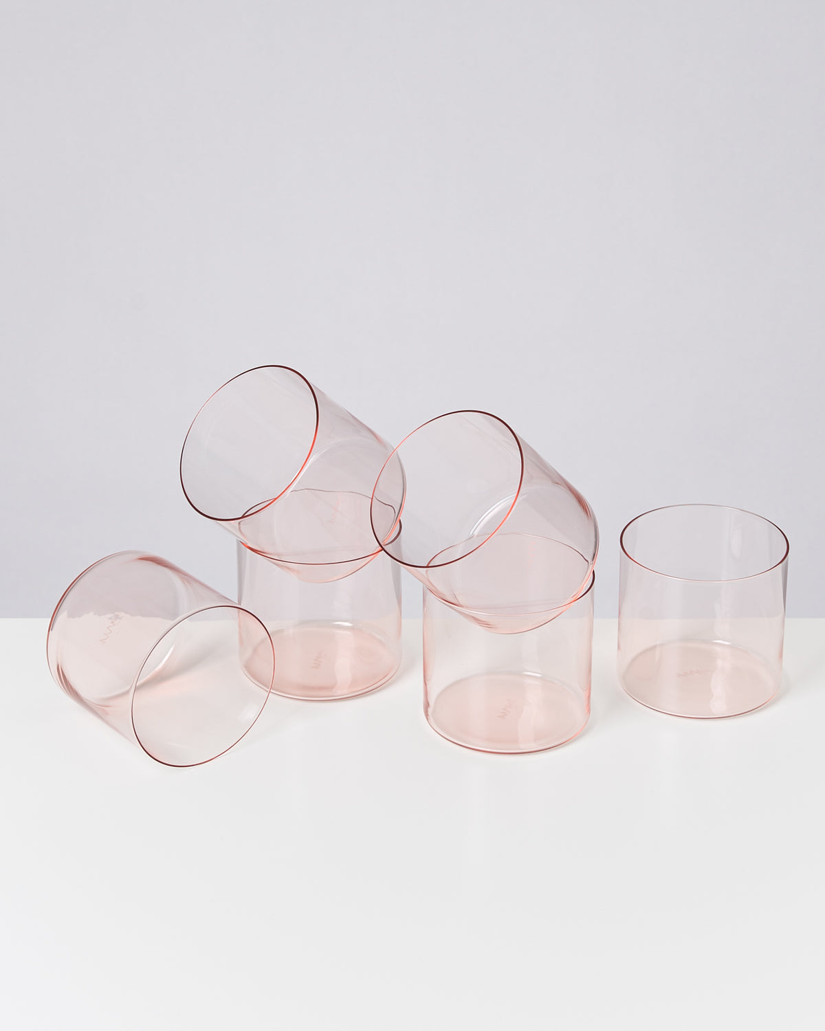 Fünf durchsichtige, rosa Tumbler-Gläser stehen und liegen auf einem weißen Tisch vor einem grauen Hintergrund.