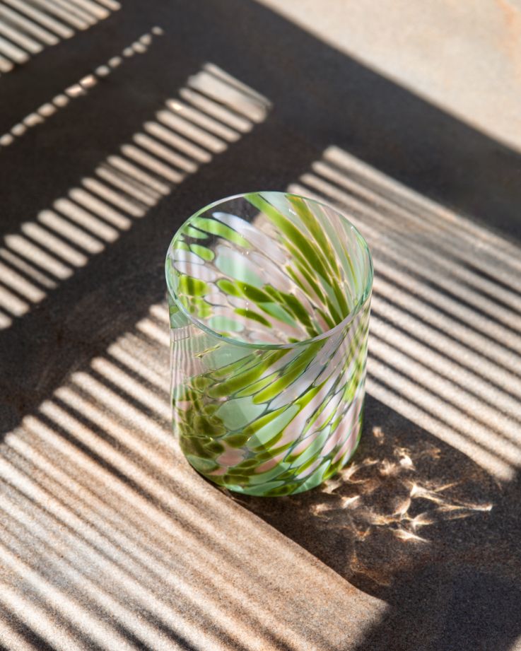 Ein Glas mit grün-weißem Wirbelmuster steht auf einem Tisch, durchzogen von Licht- und Schattenschleiern.