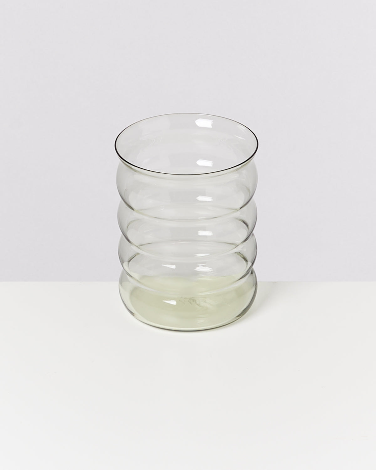 Ein klarer, wellenförmiger Glasbecher steht auf einem weißen Tisch vor einem neutralen Hintergrund.