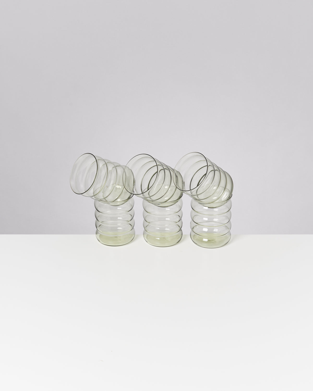Drei ineinander verschachtelte, schräg stehende, transparente Gläser auf einem weißen Tisch vor grauem Hintergrund.