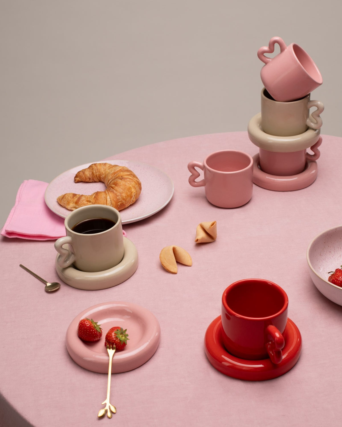 Verschiedene farbige Tassen, Croissant und Erdbeeren auf einem rosa Tisch mit elegantem Geschirr und Besteck.
