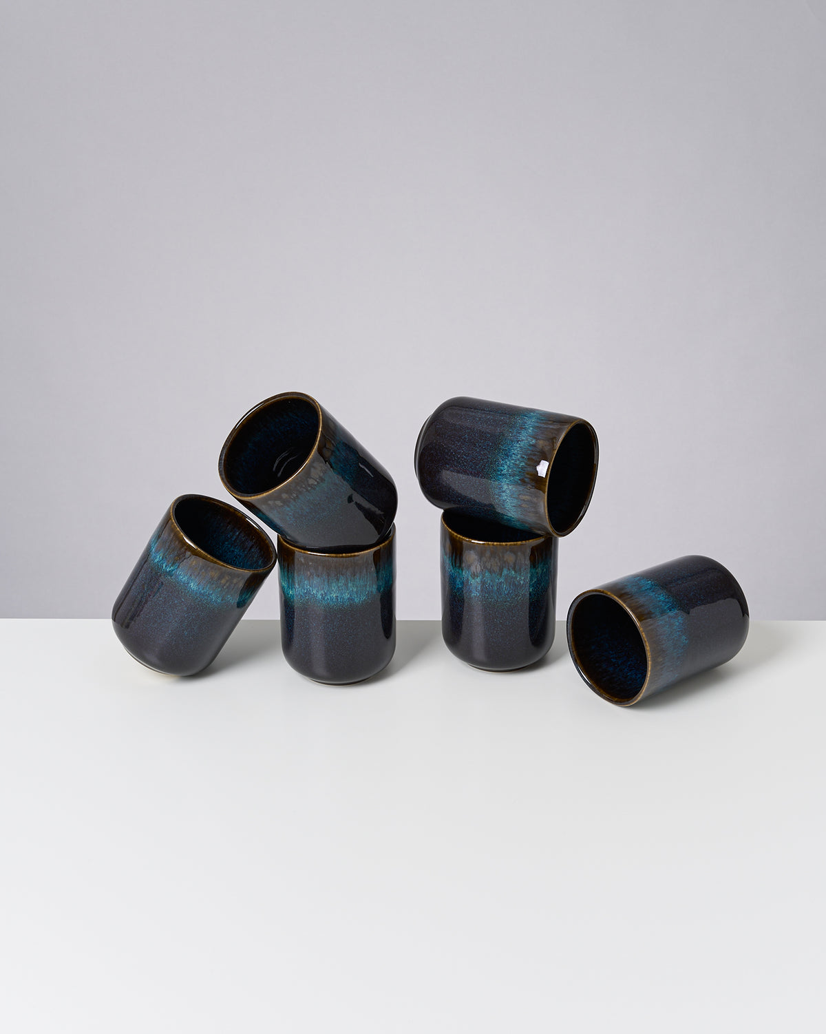 Sechs dunkelblaue Keramikbecher mit hellblauem Streifen auf einer weißen Fläche vor grauem Hintergrund.