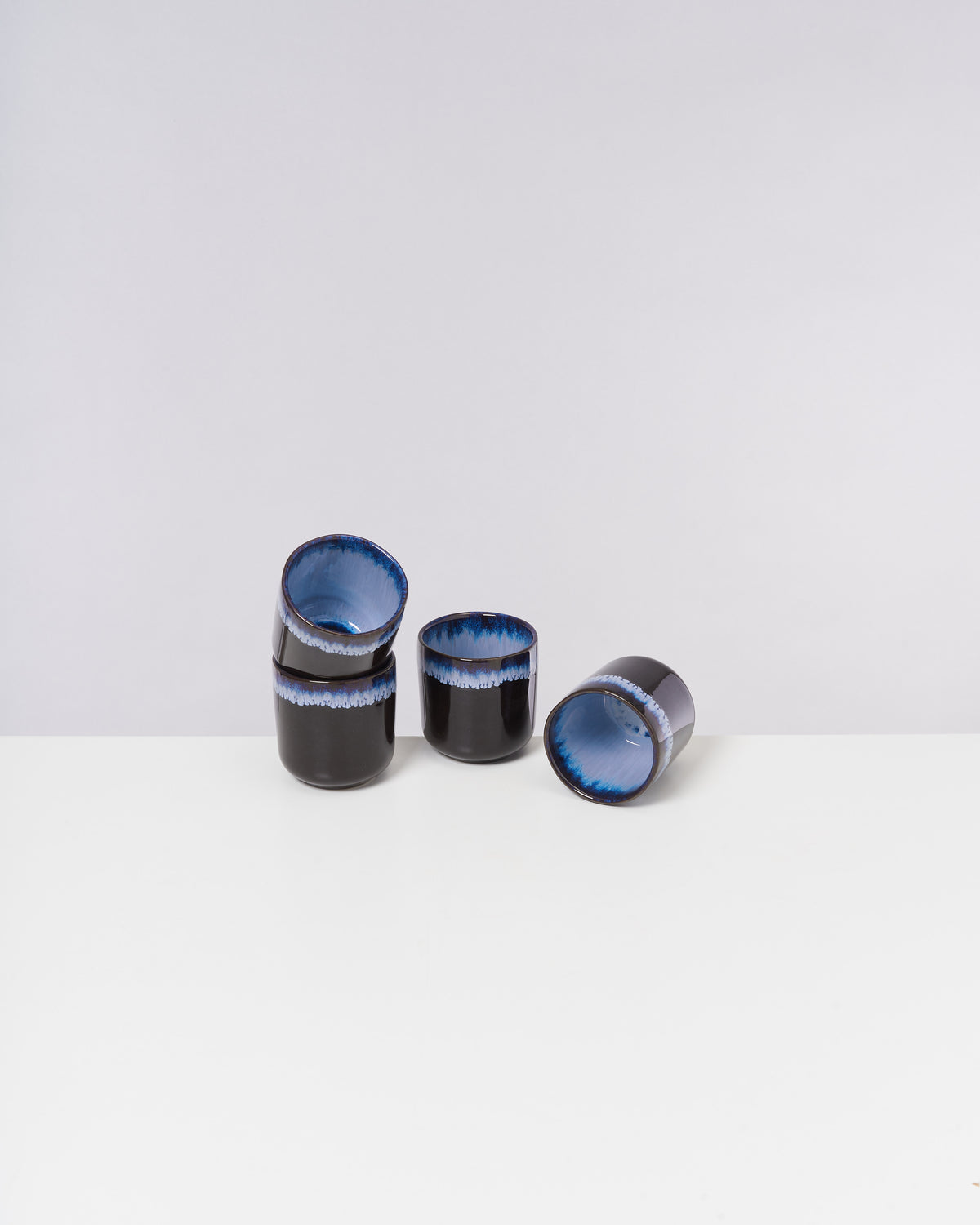 Fünf schwarze Tassen mit blauem Inneren, auf einem weißen Tisch vor grauem Hintergrund angeordnet.