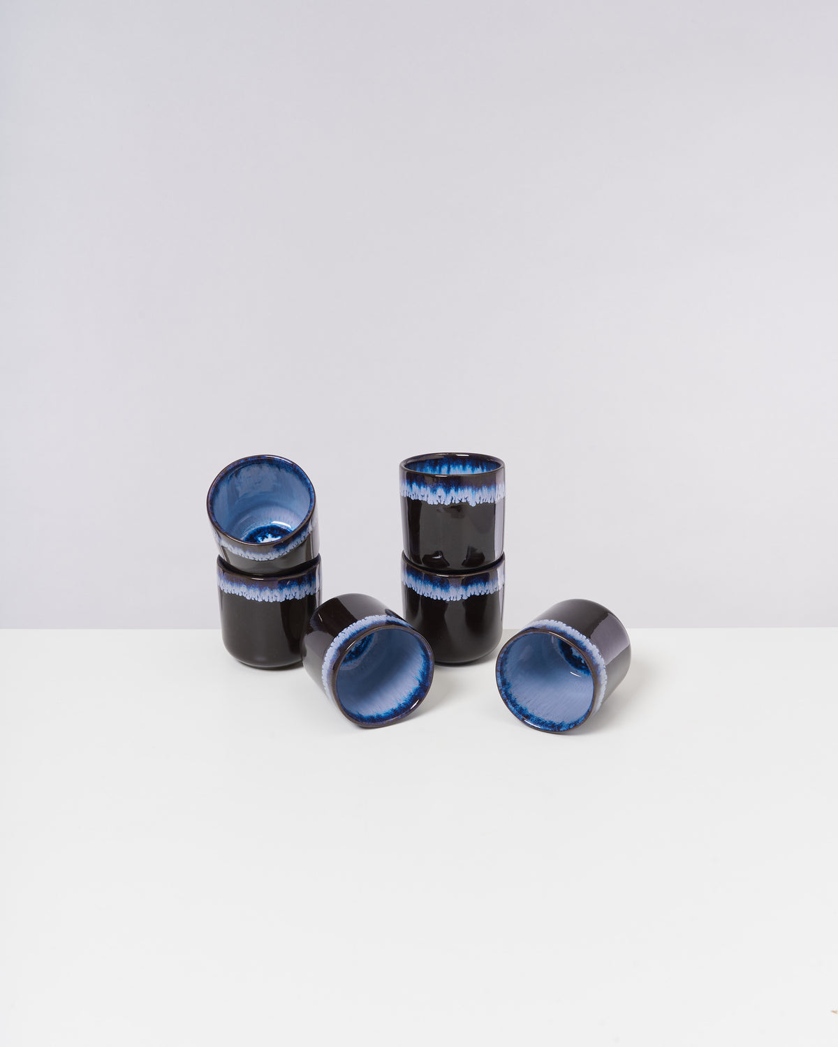 Sechs blaue und schwarze Keramiktassen, einige stehend, andere liegend, auf weißem Hintergrund.