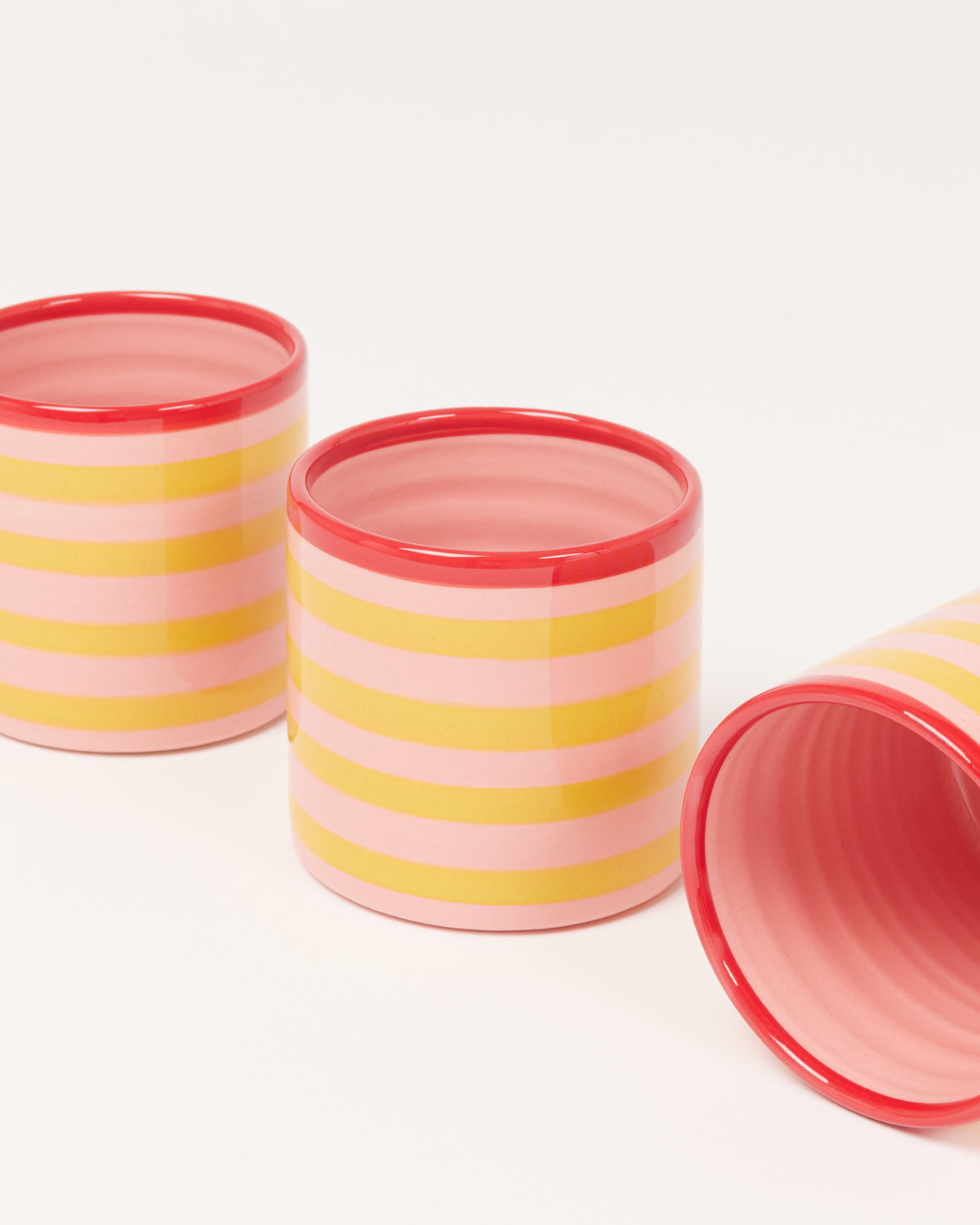 Drei Tassen mit gelb-rosa Streifen und rotem Rand auf weißem Hintergrund.