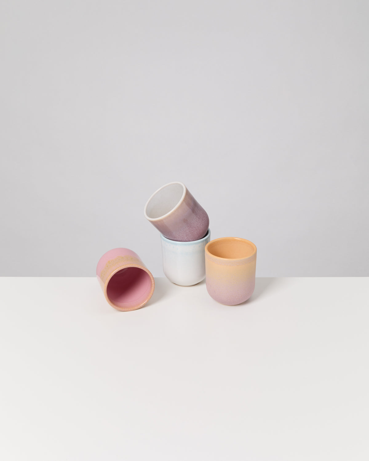 Vier bunte Keramikbecher in Pastelltönen stehen und liegen auf einem weißen Tisch vor grauem Hintergrund.