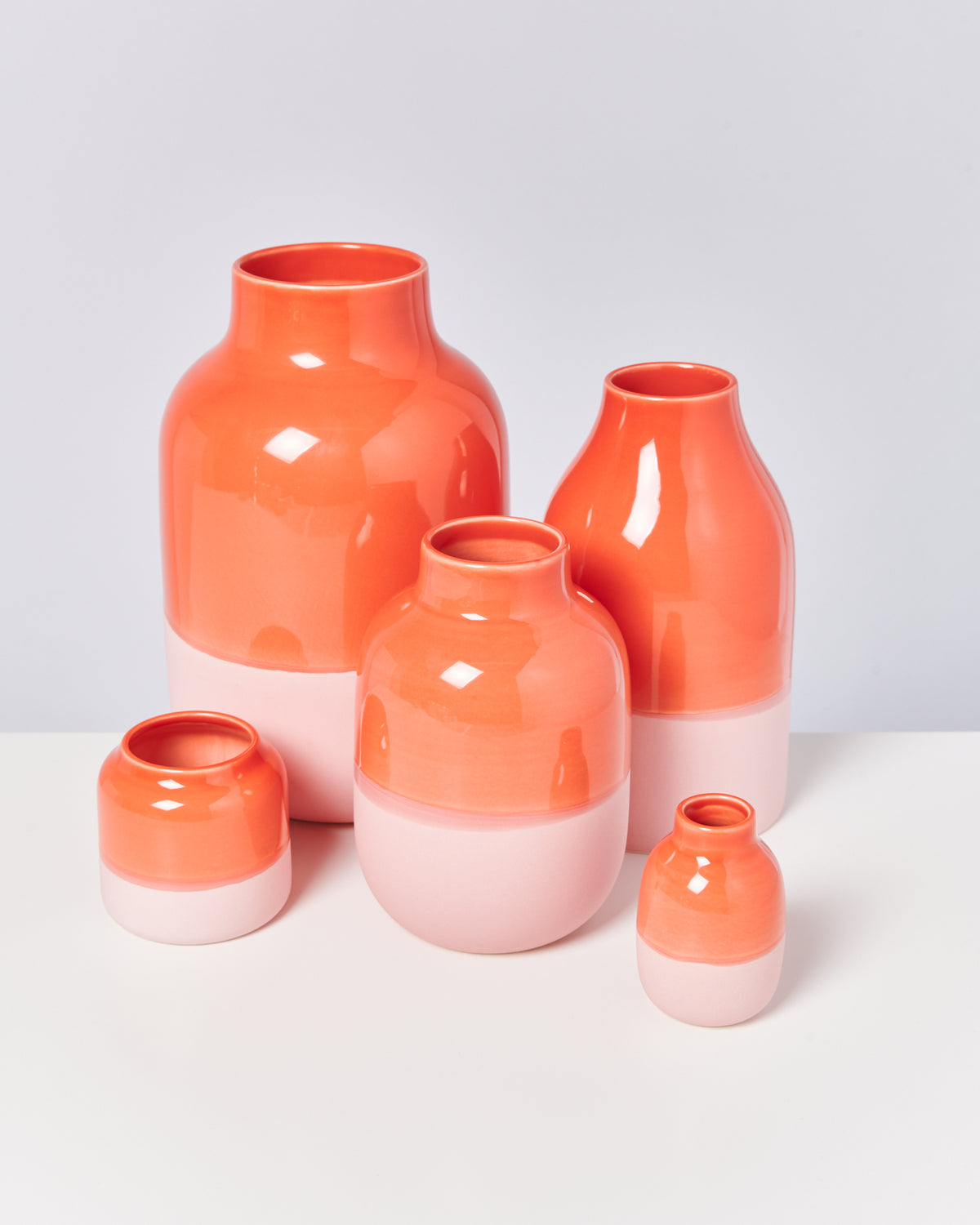 Fünf orange-rosa Vasen verschiedener Größen stehen auf einem weißen Tisch vor einem grauen Hintergrund.